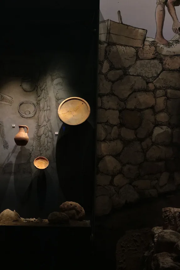 Peças de cerâmica iluminadas em vitrine ao lado de parede de pedra