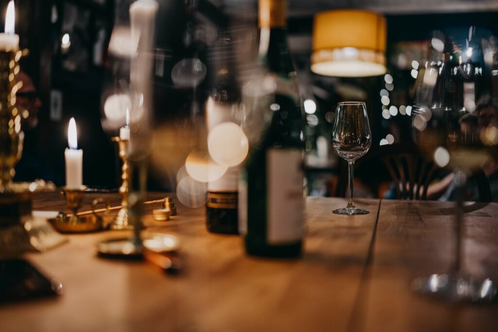 Taça de vinho sobre uma mesa de bar de madeira, com velas e garrafas desfocadas ao fundo em tons quentes, criando um ambiente aconchegante.