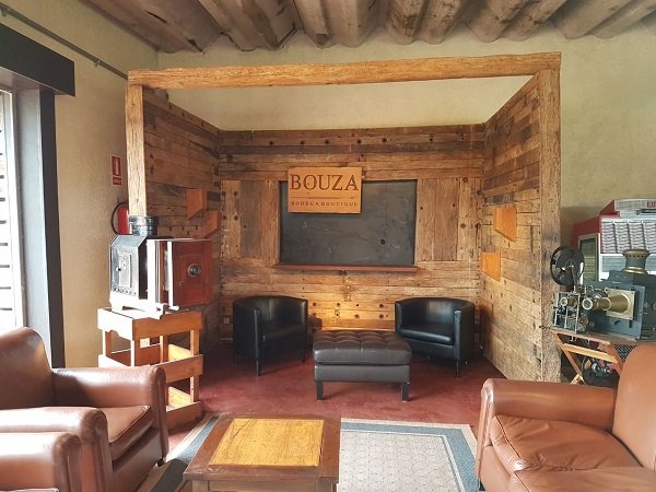 Sala de estar rústica e aconchegante com paredes revestidas de madeira, duas poltronas pretas, um pufe capitonê e uma placa "Bouza" na parede do fundo.