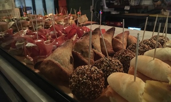 Variedade de aperitivos em tamanho de uma mordida dispostos em um expositor de vidro, incluindo bolinhos com crosta de gergelim, minissanduíches e petiscos no espeto, prontos para servir.