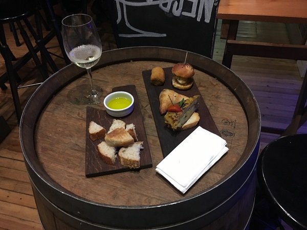 Mesa feita de barril de vinho com uma taça de vinho branco, duas tábuas de madeira com tapas (cubos de pão com azeite) e mini sanduíches, além de uma pilha de guardanapos, em um ambiente de restaurante.