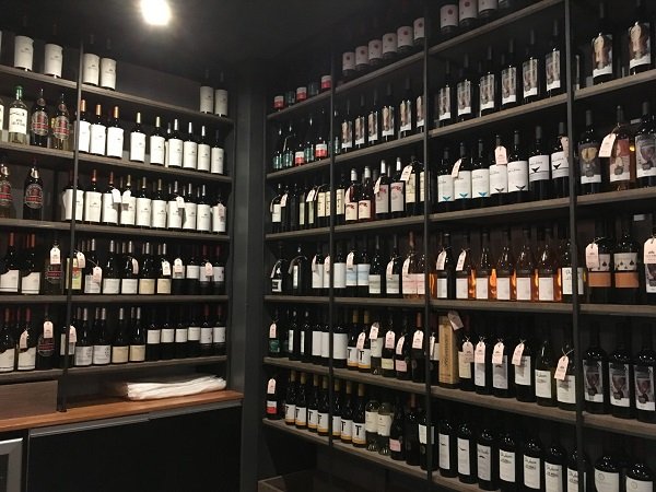 Prateleiras repletas de garrafas de vinho em uma loja de vinhos ou adega de iluminação tênue, dispostas em longas fileiras ao longo de estantes de metal escuro.