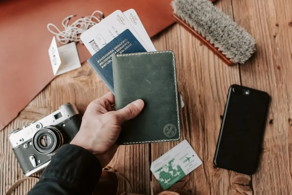 Mao segurando um passaporte ao lado de camera, cartoes e celular sobre a mesa