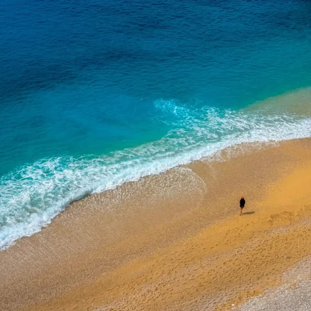 Praia de areia clara com mar calmo e uma pessoa caminhando a beira da agua