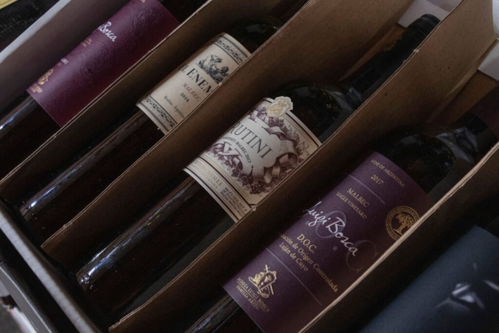 Garrafas de vinho acomodadas em uma caixa de madeira, com rótulos em tons de roxo e creme visíveis nas garrafas.
