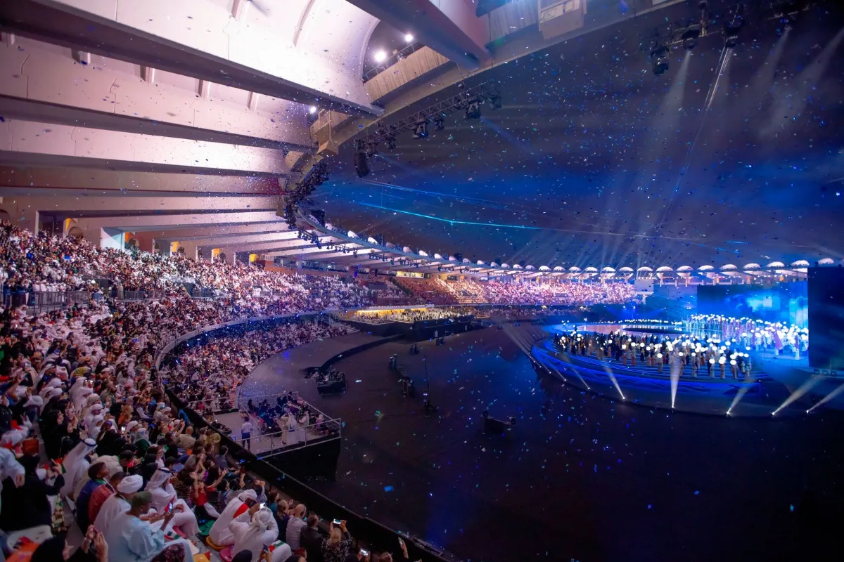 Publico de um show em uma arena coberta, com o palco iluminado ao fundo
